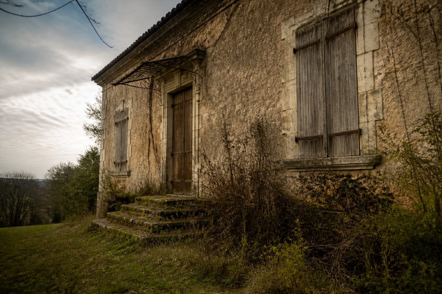Façade d’une maison ancienne abandonnée en zone rurale française avec volets fermés et végétation envahissante